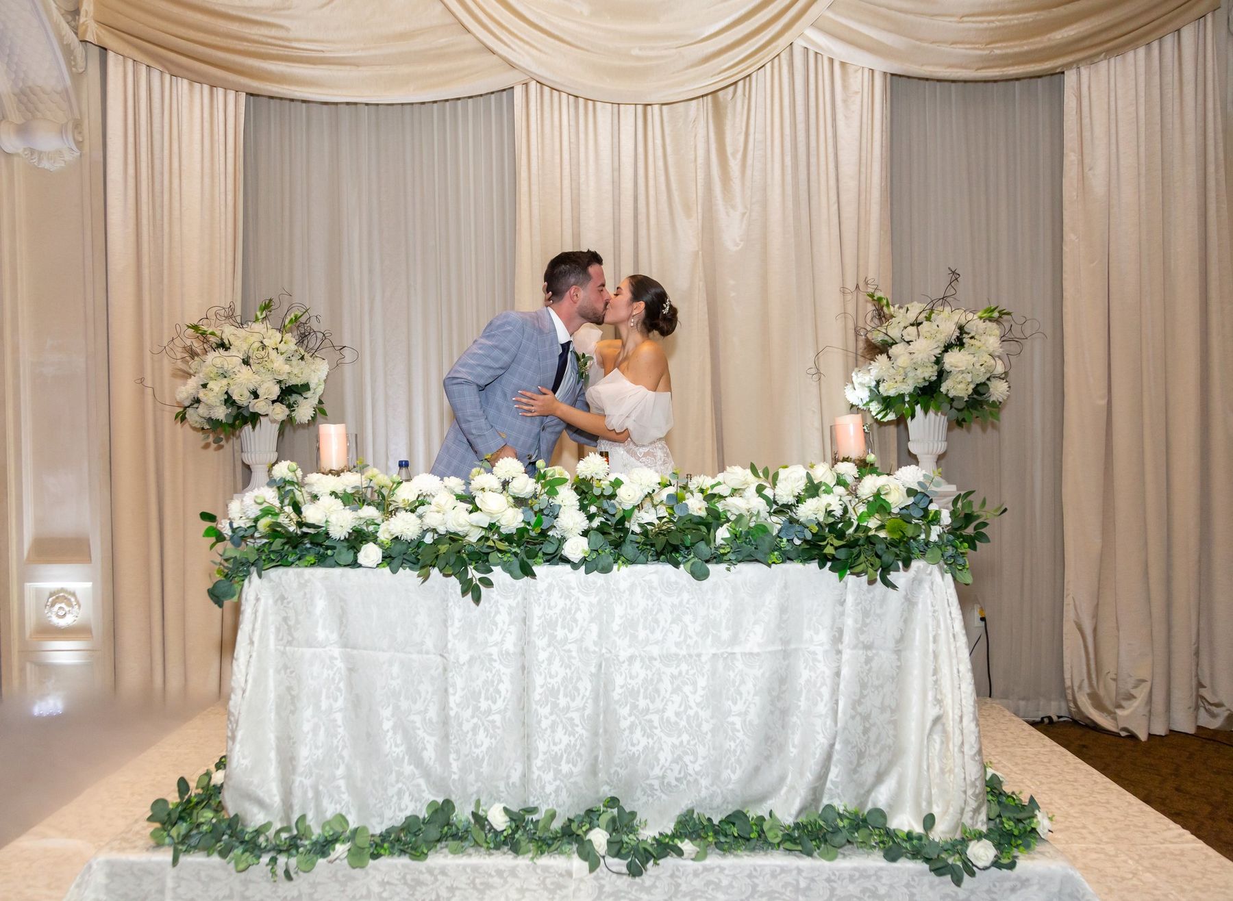 A couple shares a kiss behind their sweetheart table under champagne drapery